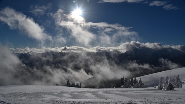 A snowy landscape with rising clouds and distant peaks.