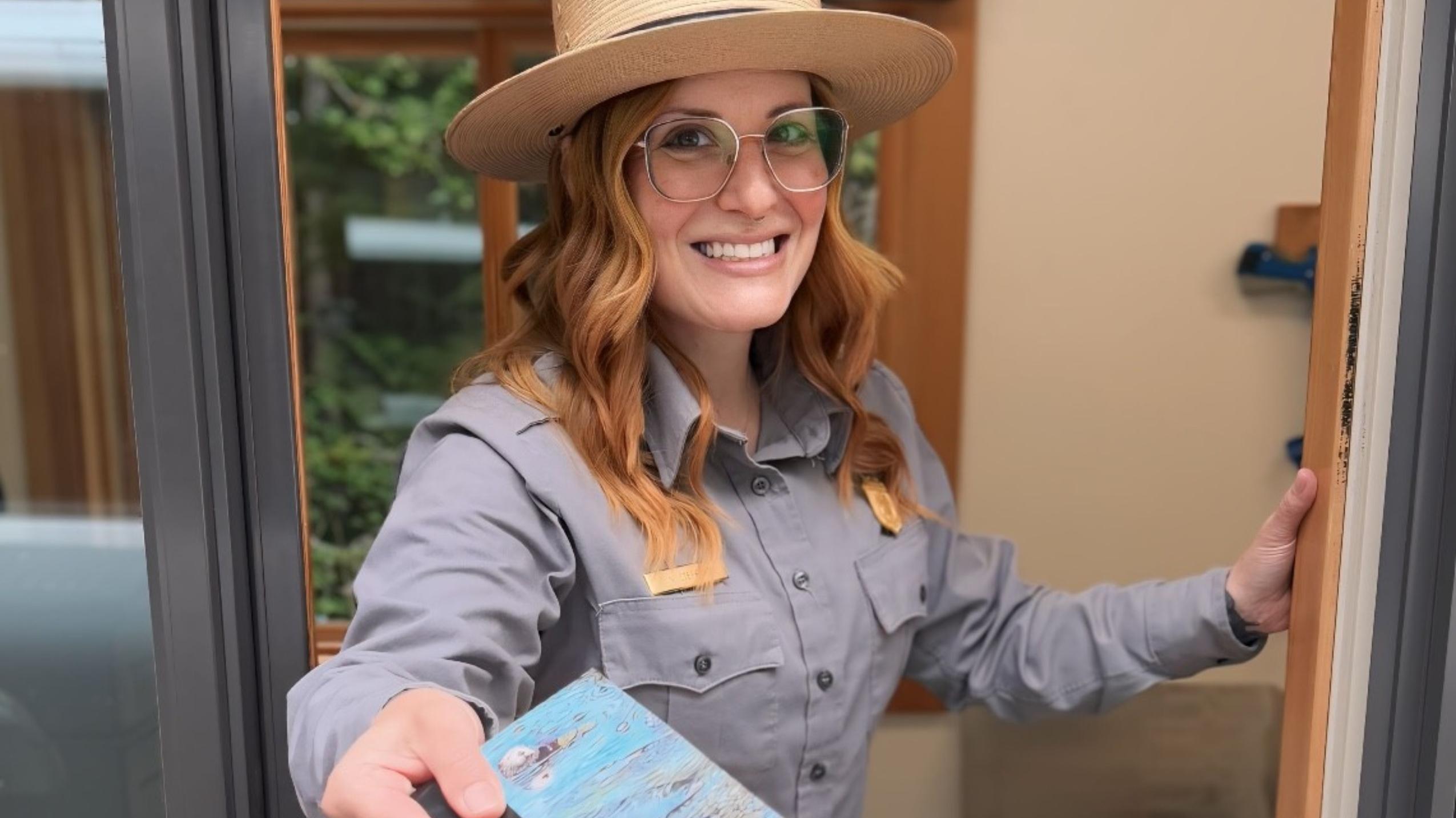 A smiling woman with wavy red hair and glasses in a national park uniform extends a map toward you
