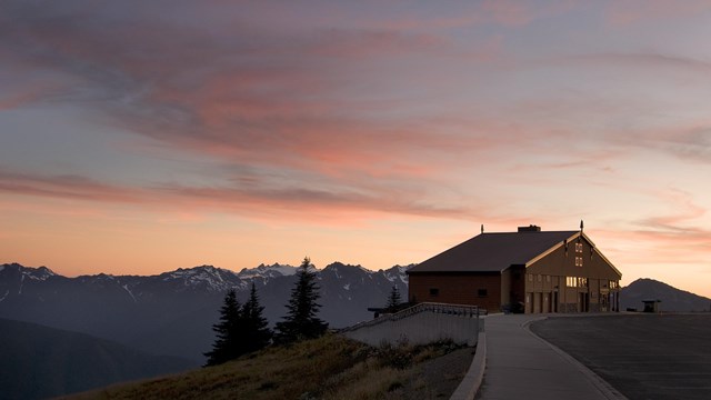 A building on a hillside with a mountain vista beyond at sunset.