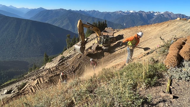A large piece of machinery excavates a slope with the Olympic Mountains in the background.