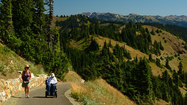 A man in a wheelchair and woman walking on a paved trail on a hillside.