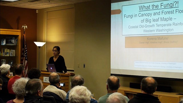 A woman presenting a slideshow to a crowd, titled "What the Fungi?!"