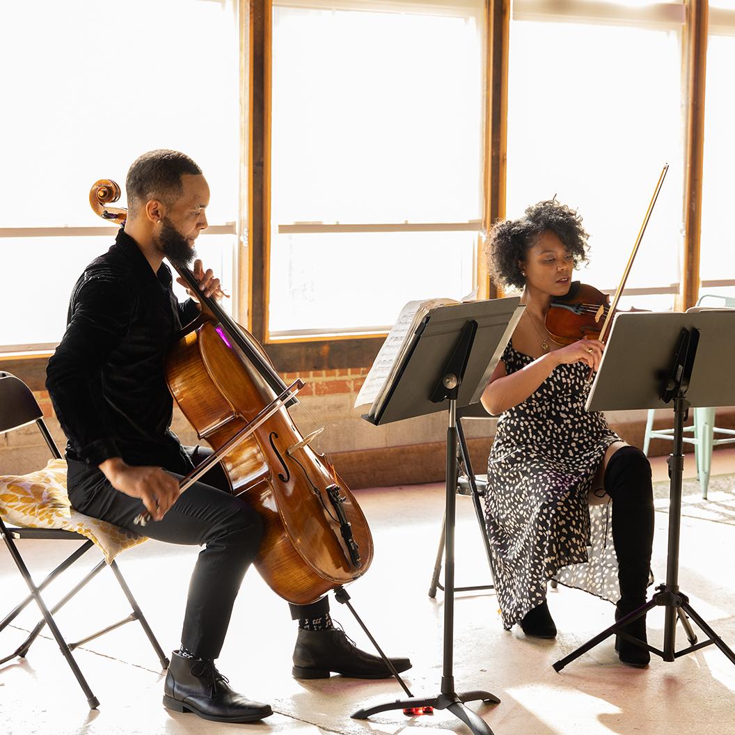 A Black man with a cello and a Black woman with a violin in a large, sunlit room.