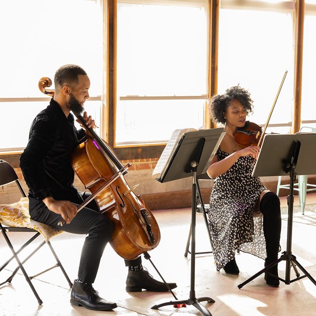 A Black man with a cello and a Black woman with a violin in a large, sunlit room.