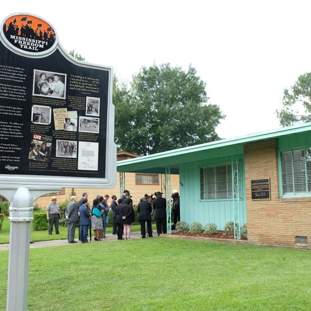 A placard for the Mississippi Freedom trail outside a single story house, with people gathered.