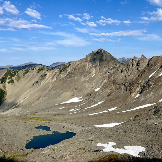 Sweeping blue skies dotted with clouds above mountain peaks and rocky glacial rubble, and a pond