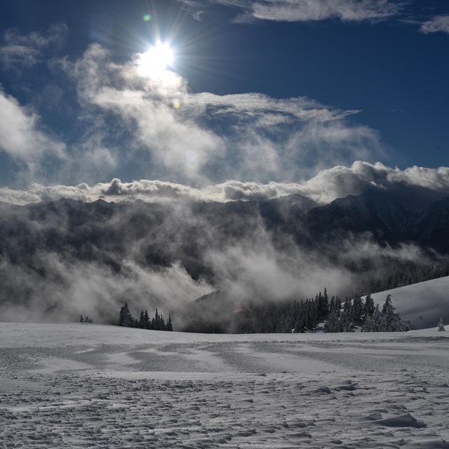 A snowy landscape with rising clouds and distant peaks.
