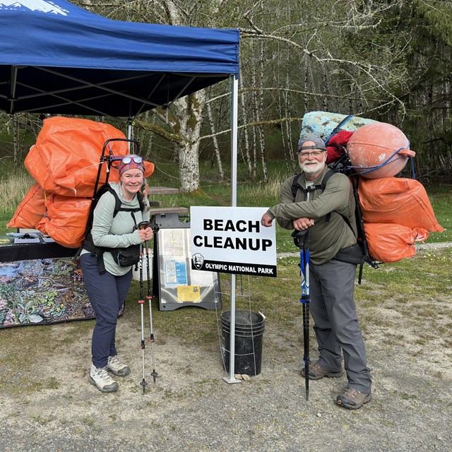 Two volunteers carrying trash from the beach on their back next to a beach cleanup sign.
