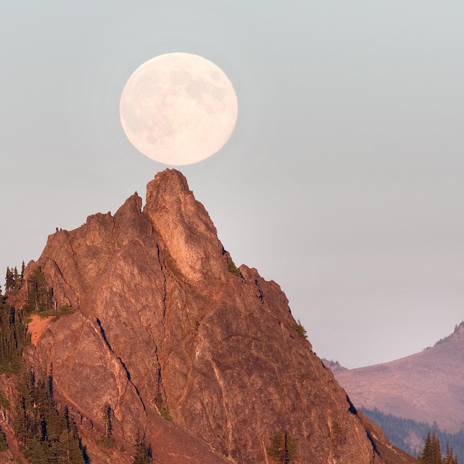 A huge, full moon hangs just above the summit of a rocky mountain peak.