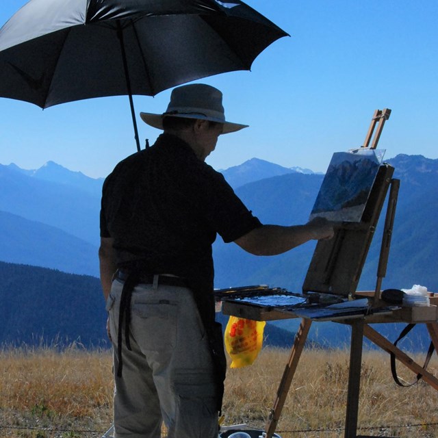 A man standing with a sunhat on is painting the mountains and trees in the background. 