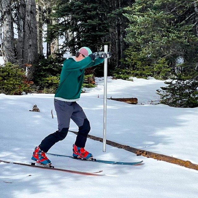A young man in winter gear holds up a metal tube, as if about to plunge it into the snow.