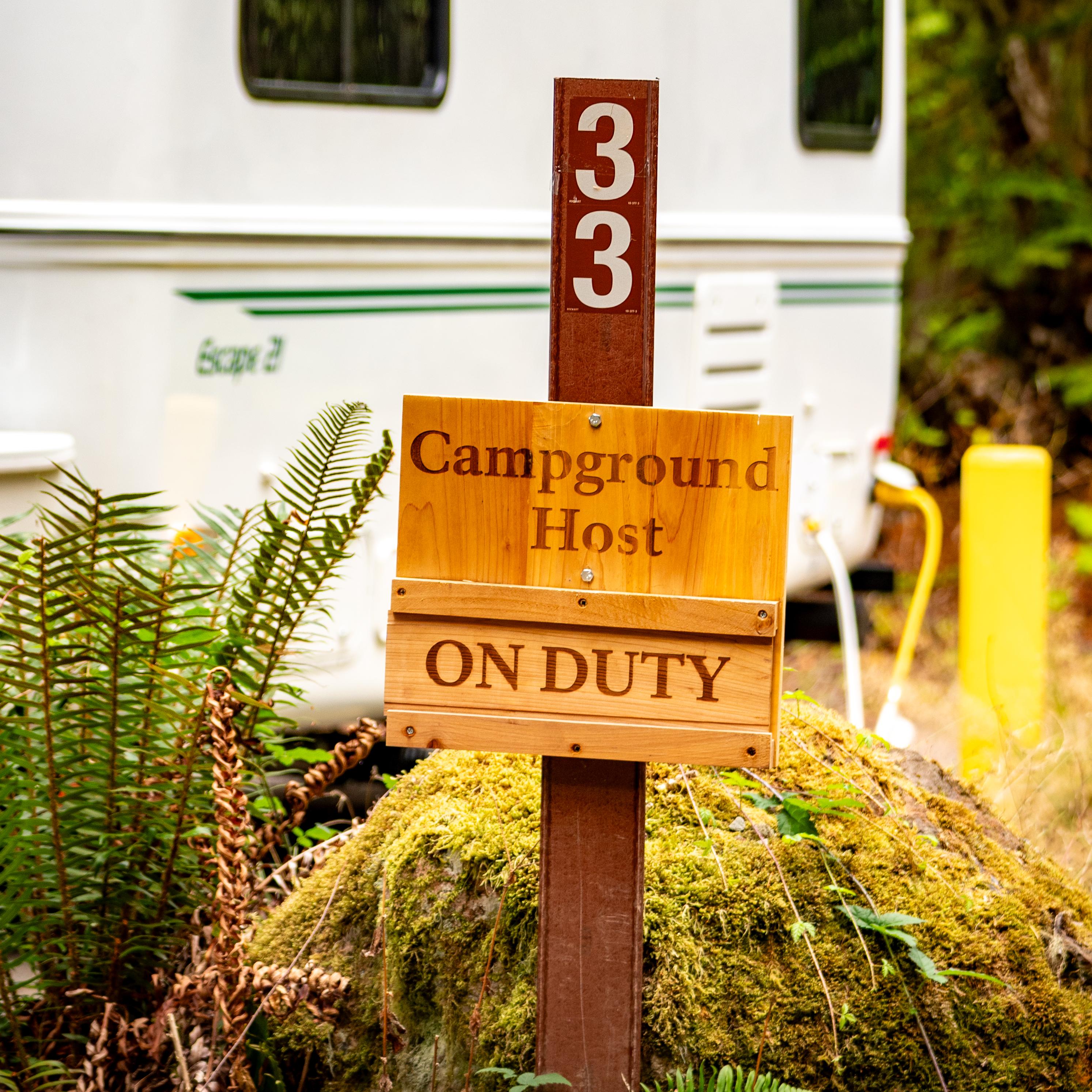 A wooden sign reads Campground Host On Duty. Beyond there is an RV in parked in a mossy forest.