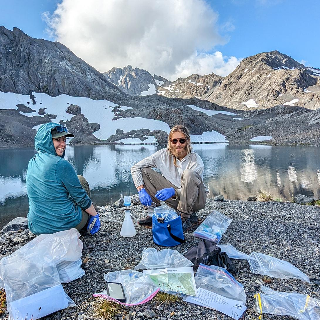 Two scientists, smiling in hiking clothes and latex gloves, sit on the shore of a mountain lake. Map