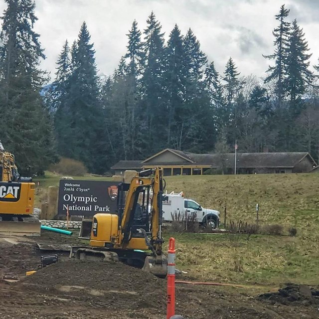 Construction equipment partially obscures a park entrance sign and building among tall trees.
