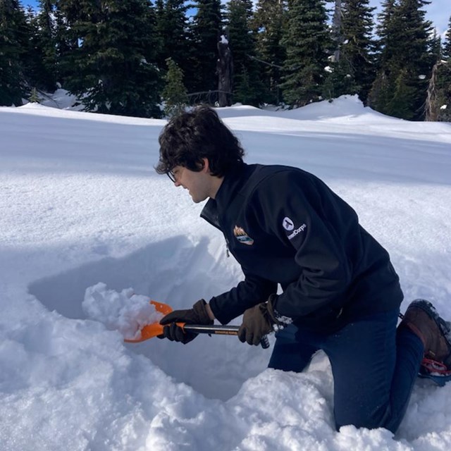 A young man with wavy dark hair and glasses kneels in the now, using a small shovel to dig a trench