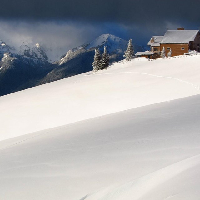 A lodge building perches on a snowy hillside, with dramatic snowy mountain peaks beyond.