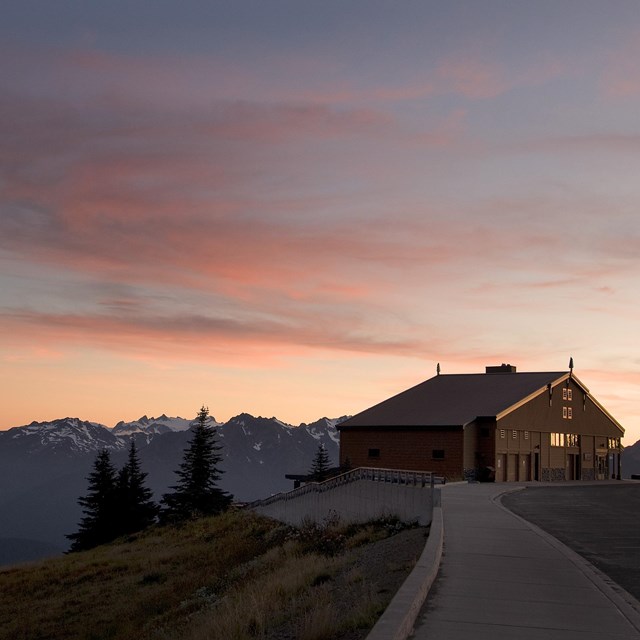 A building on a hillside with a mountain vista beyond at sunset.