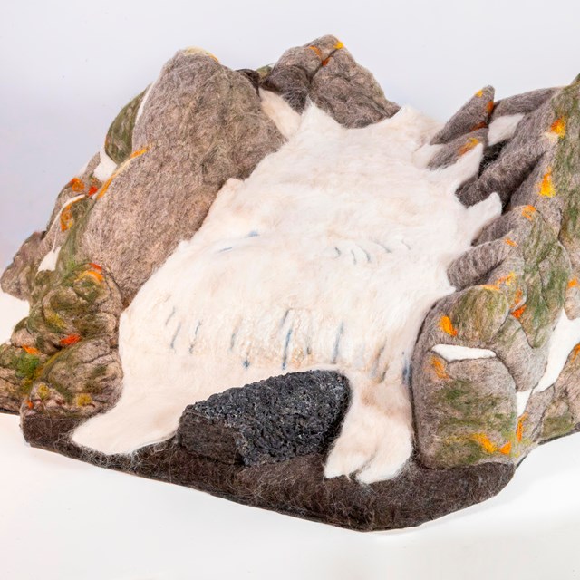 A mountain glacier sculpted from wool felt. The white ice of the glacier flows between grey “rock” r