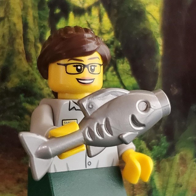 A LEGO minifigure with glasses and a ponytail holds a large fish in her hand.