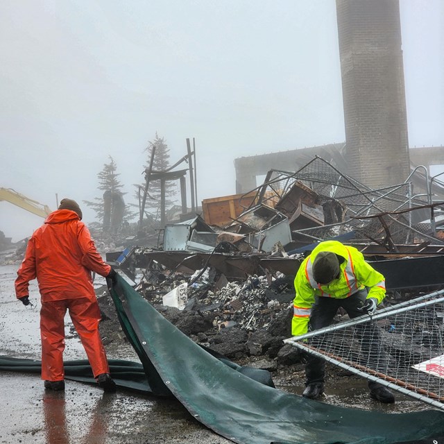 Two workers in hatss and orange and neon yellow rain gear move a tarp and tent near burned wreckage.