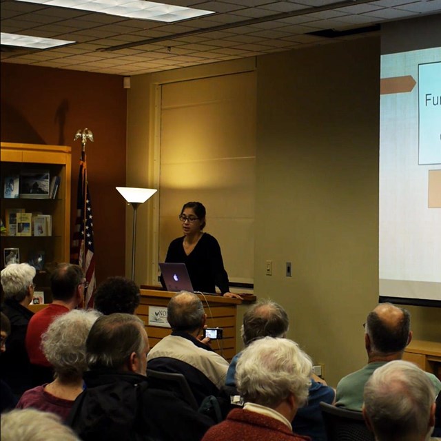 A woman presenting a slideshow to a crowd, titled 