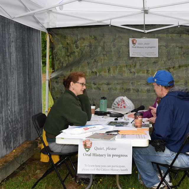 A historian and event visitor sit across from each other at a table under an outdoor tent and talk.