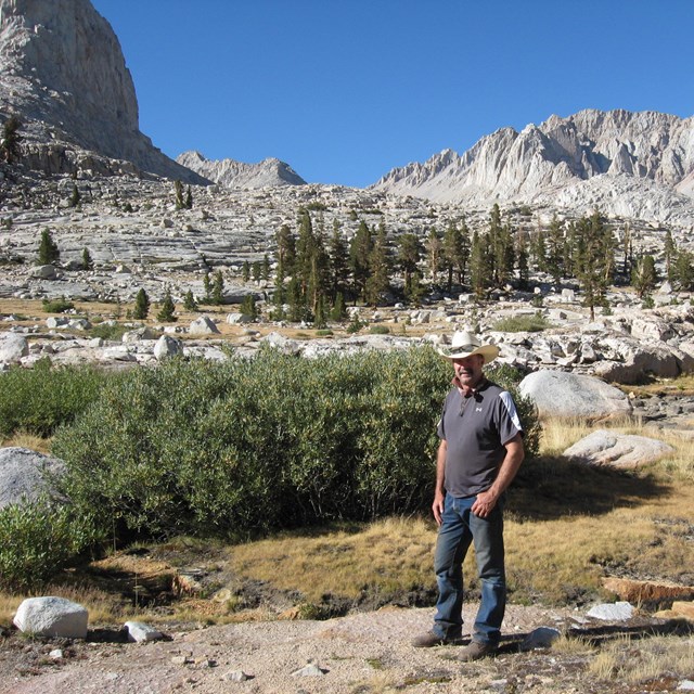 J.D. Swed poses, in cowboy hat, amid a rocky landscape of low grass, conifers, and mountains.