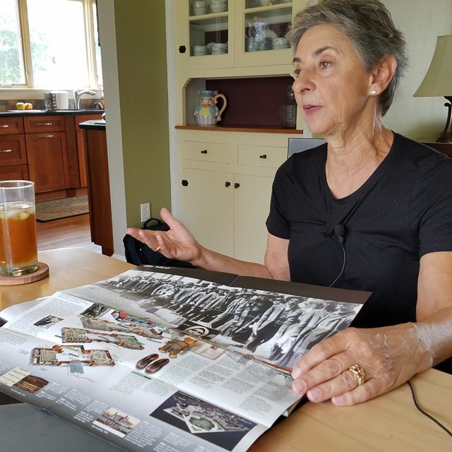 A women, seated at a dining room table, gestures over the park brochure in front of her.