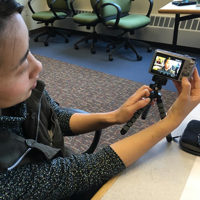 A interviewer holds up a camera mounted on a tabletop tripod to show the display of a recorded video