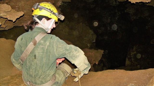 Off trail caver in jumpsuit and hard hat sitting on cave floor and holding a rope.