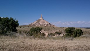 A pointed bluff landmark sticks out above a flat valley with large green shrubs.