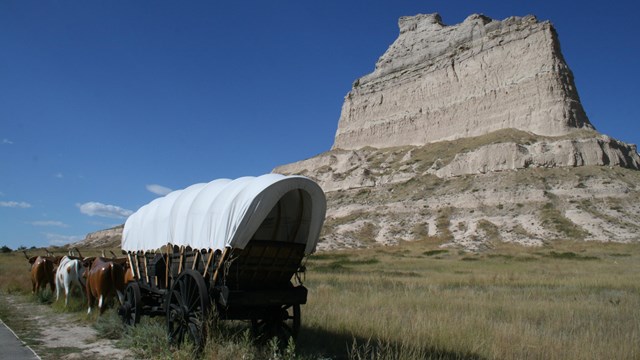 A large covered wagon in front of a towering sandstone bluff.