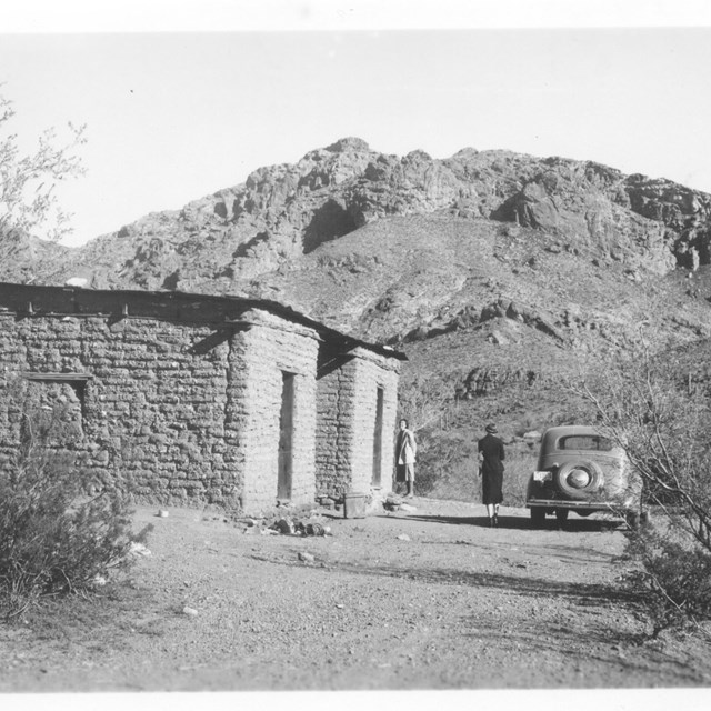 An old car next to an adobe structure, with family present