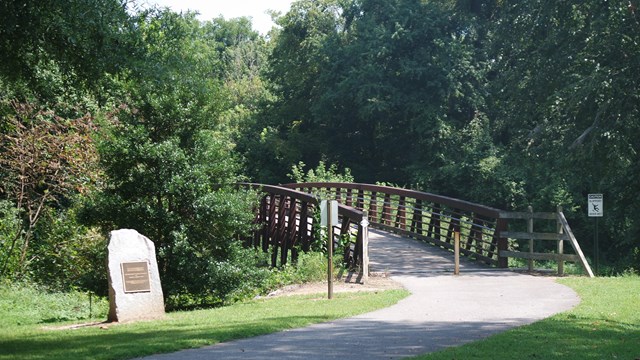 A paved trail leads to a metal bridge crossing a river with wooded banks, passing a stone marker.