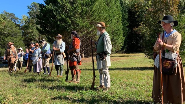 Line of people dressed as frontier soldiers stand with rifles.