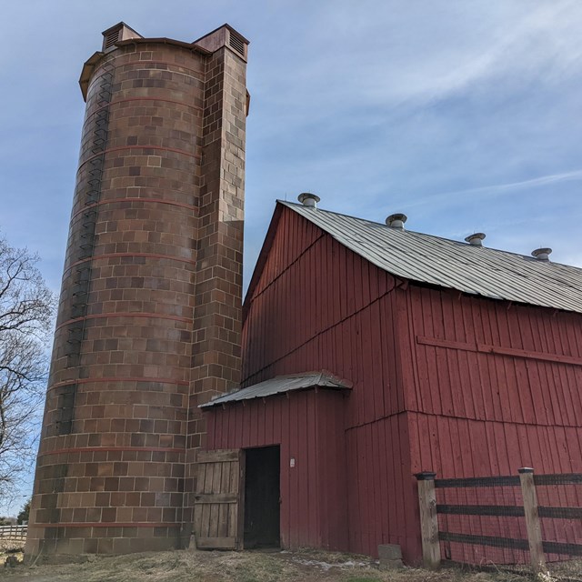 A brown tile silo attached to a red dairy barn