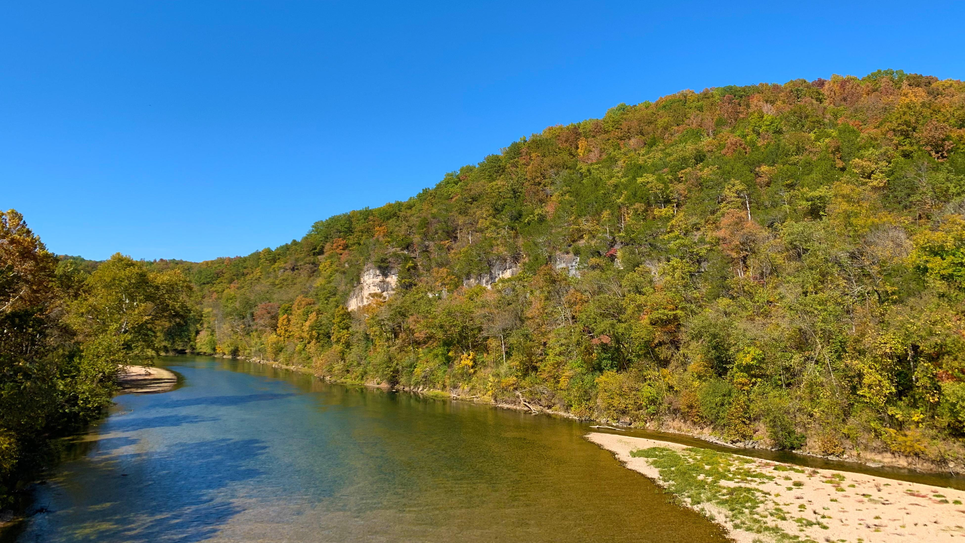A river scene of a clear stream flowing through rolling hills with fall colors in the trees
