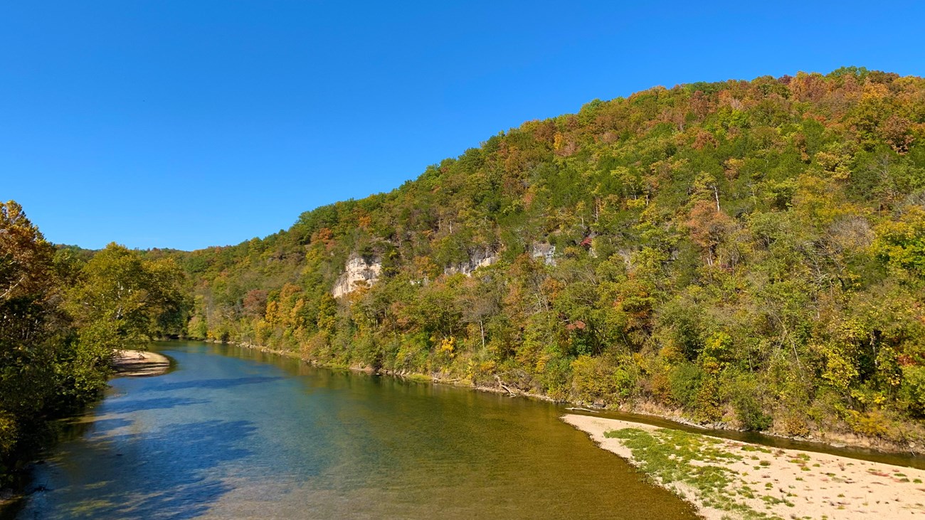 A river scene of a clear stream flowing through rolling hills with fall colors in the trees