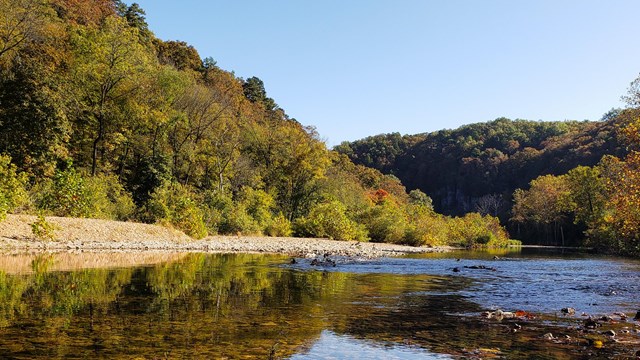 A wide river flows between rocky bluffs and wooded hills. There is beautiful fall color all around.