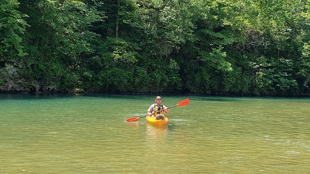 A park ranger floats in a river. Trees cover the background.