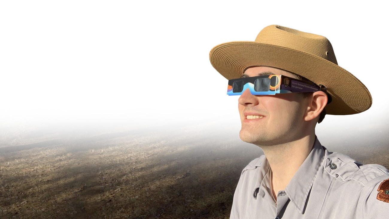 A park ranger wears eclipse glasses and looks towards the sun.