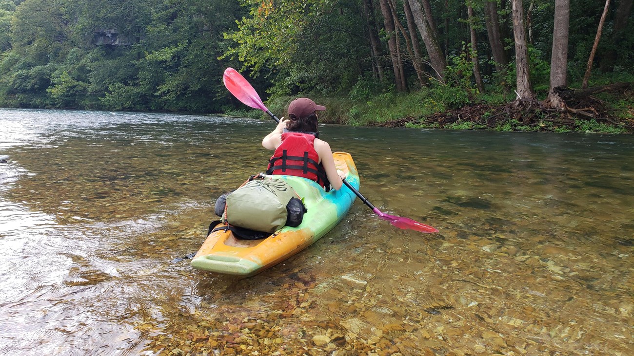 A multi-colored kayak with paddler up close on a shallow clear stream