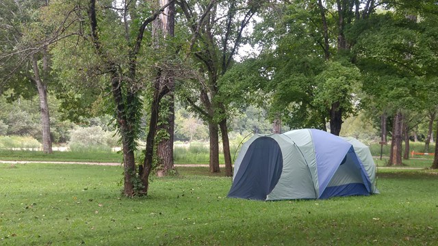 A tent sits in a field with a few trees nearby. There is a river in the background.