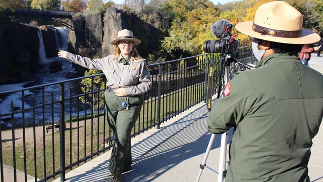 A female ranger gestures toward the Great Falls while filmed by male ranger.