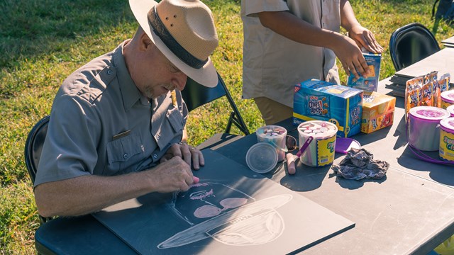 A park ranger draws himself on a blackboard slate with chalk at a table outside