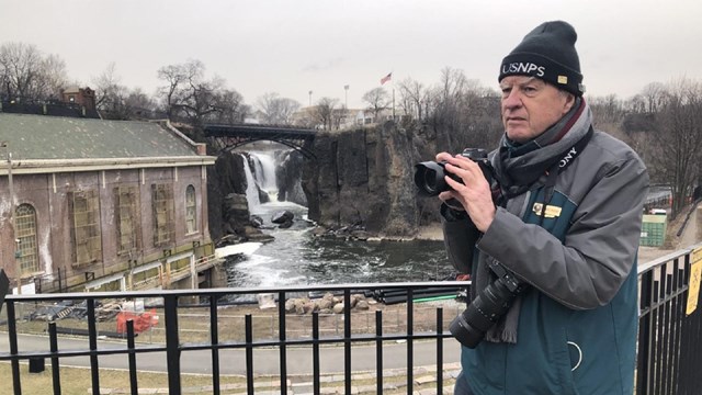 A volunteer in parks with a camera standing before a waterfall framed by a bridge
