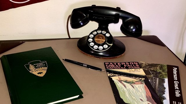 Brochures for Paterson Great Falls NHP sit by a rotary phone, a red Paterson pennant, & a notebook