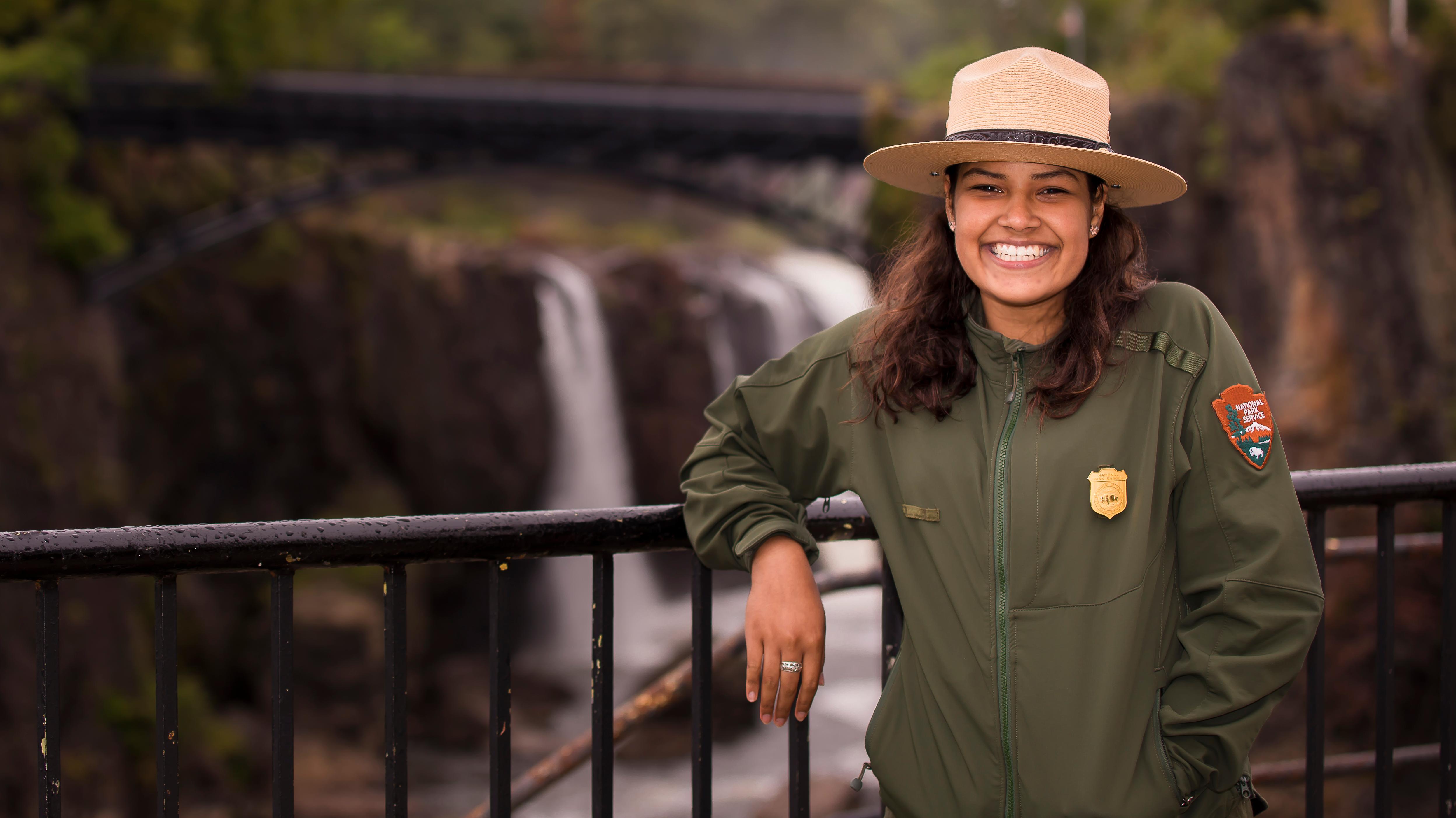A smiling park ranger leans on a railing in front of the 77 ft. Great Falls