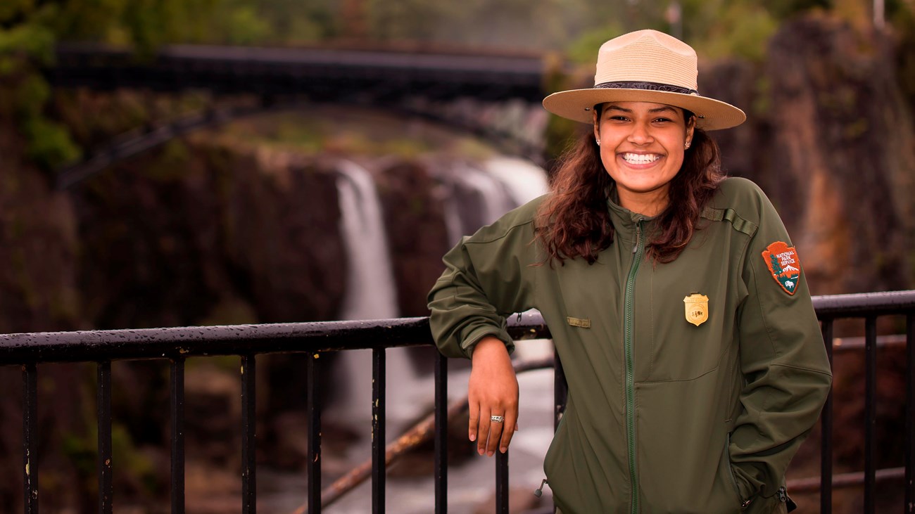 A smiling park ranger leans on a railing in front of the 77 ft. Great Falls