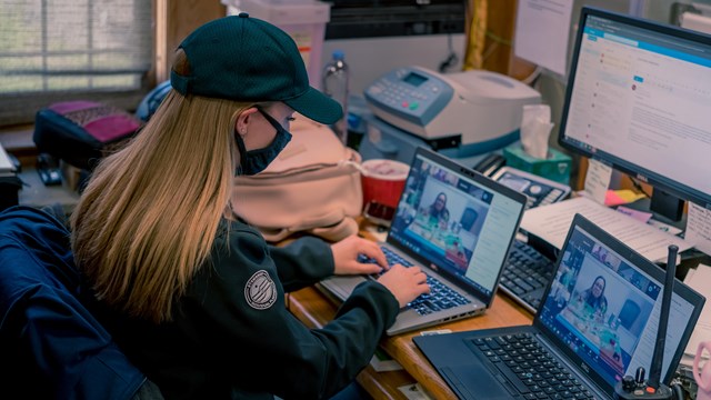 A woman working on multiple computers & monitors, a Park Ranger visible on some screens 